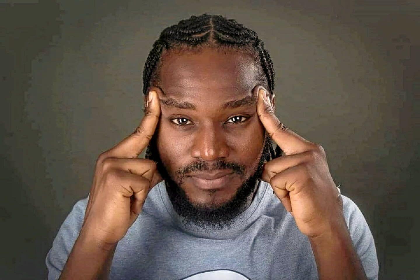 Man with braided hair and beard touching his temples, wearing a gray shirt.